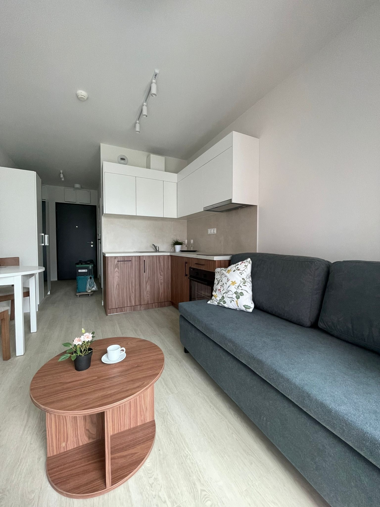 Jasne wnętrze mieszkania po remoncie: szary narożnik, drewniany stolik kawowy z kwiatami i filiżanką, zabudowa kuchenna w bieli i drewnie. Widok na odnowioną przestrzeń.