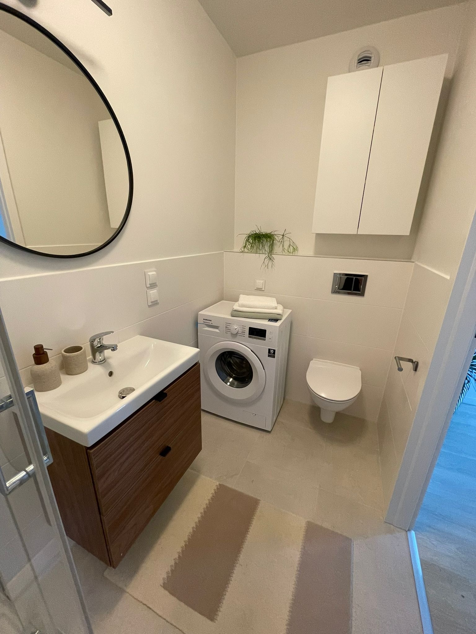 Nowoczesna łazienka w Chojniczkach z drewnianą szafką pod umywalką, pralką i wiszącą toaletą. Minimalistyczny design, jasne płytki i okrągłe lustro.