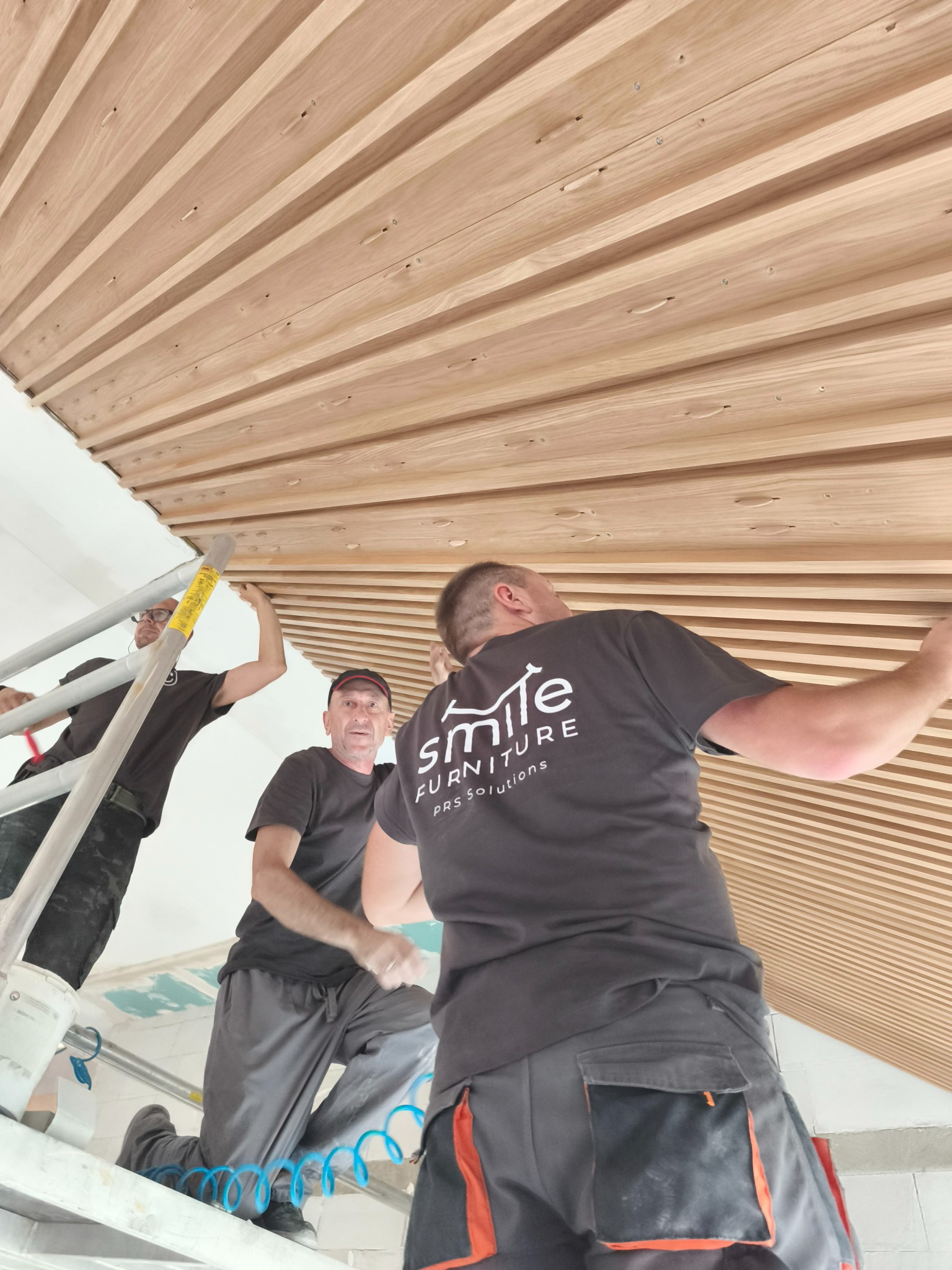 Montaż drewnianego sufitu lamelowego przez trzech monterów na rusztowaniu. Widoczne narzędzia i logo firmy na koszulce jednego z nich. Nietypowy układ lameli.