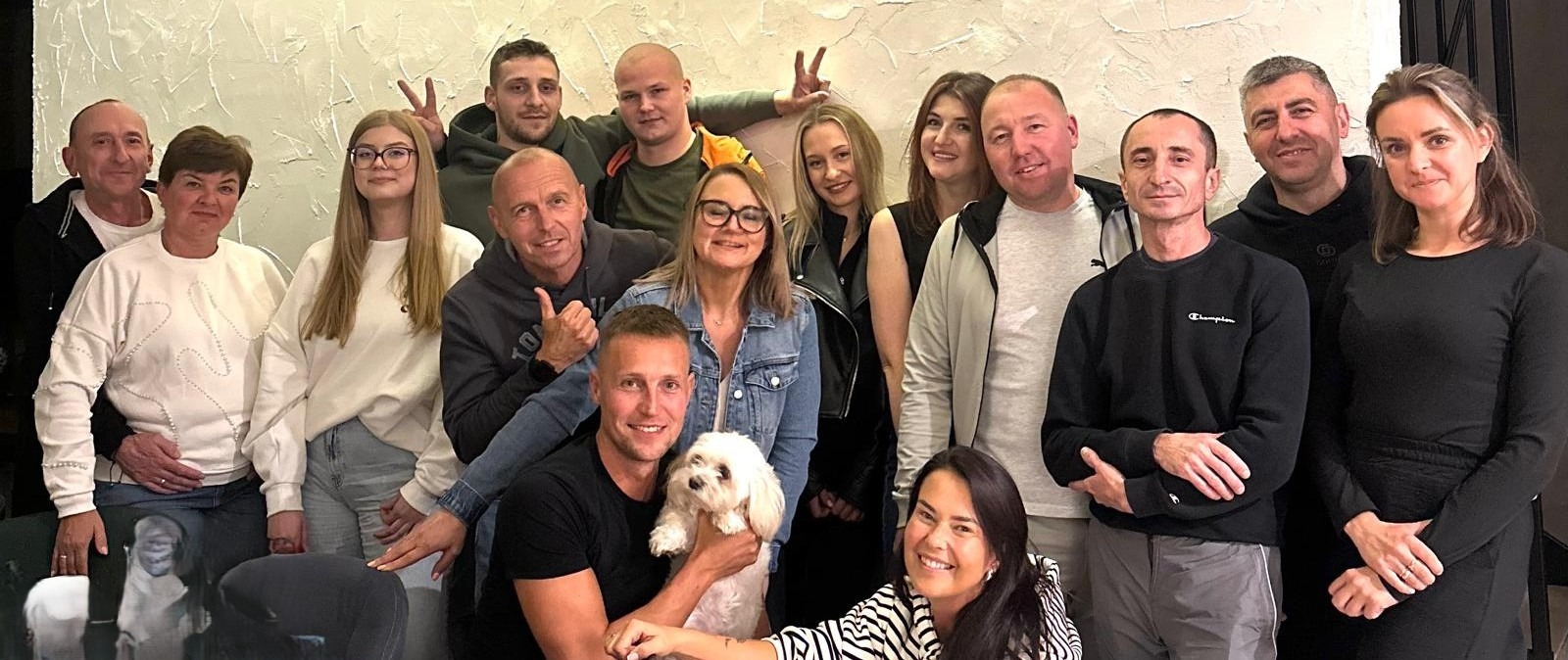 Grupowe zdjęcie zespołu w swobodnej atmosferze, uśmiechnięte twarze, różnorodne style ubioru, jasne tło z teksturą, mężczyzna trzymający małego psa.