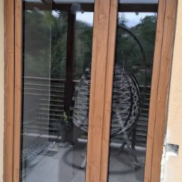 Brązowe okno PCV z widocznymi śladami montażu, pianką uszczelniającą i częściowo wykończonym tłem. Widok na zewnątrz przez szybę.