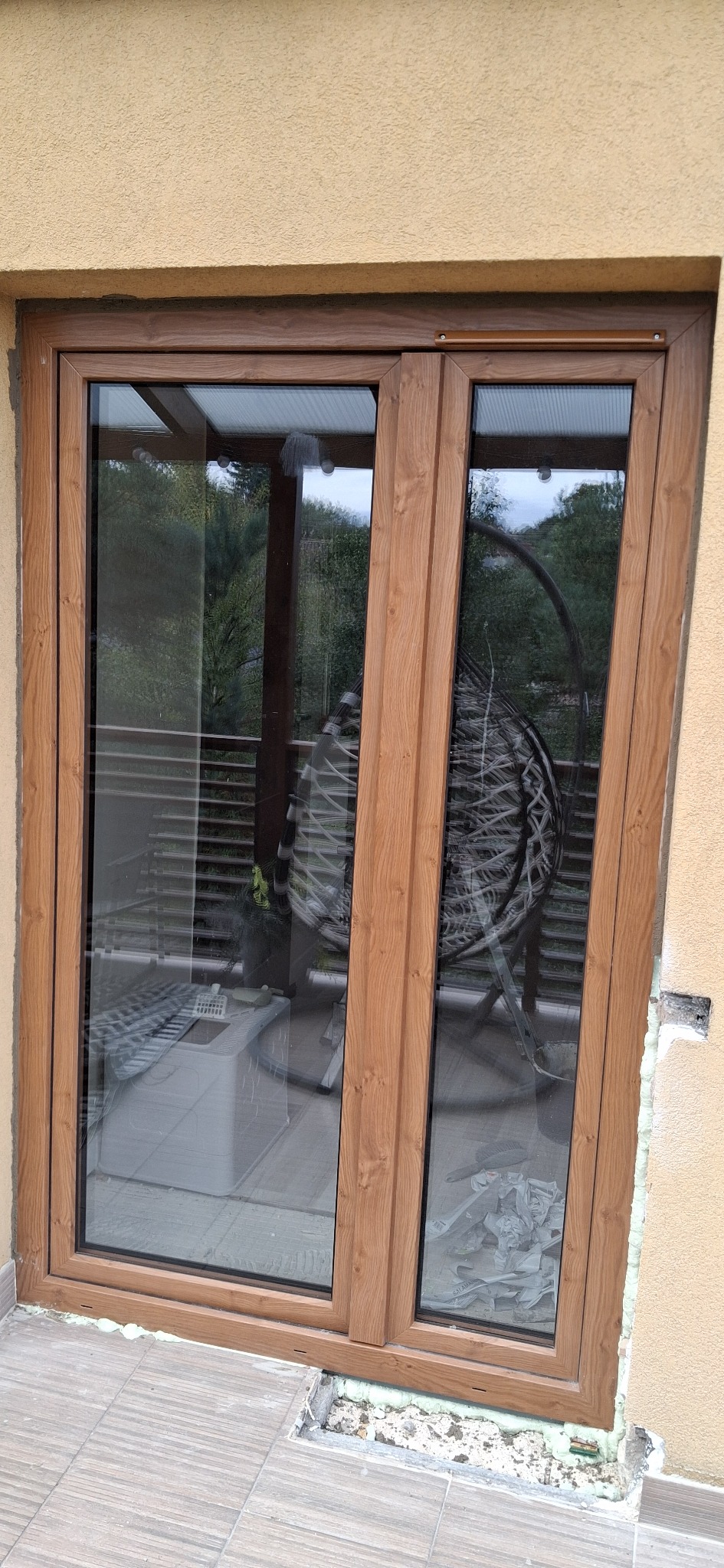 Brązowe okno PCV z widocznymi śladami montażu, pianką uszczelniającą i częściowo wykończonym tłem. Widok na zewnątrz przez szybę.