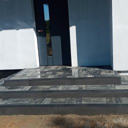 Dorian Sakowski - Schody wejściowe z kostki brukowej w odcieniach szarości prowadzące do nowoczesnego domu z ciemnymi drzwiami. Widoczne detale wykończenia i otoczenie.