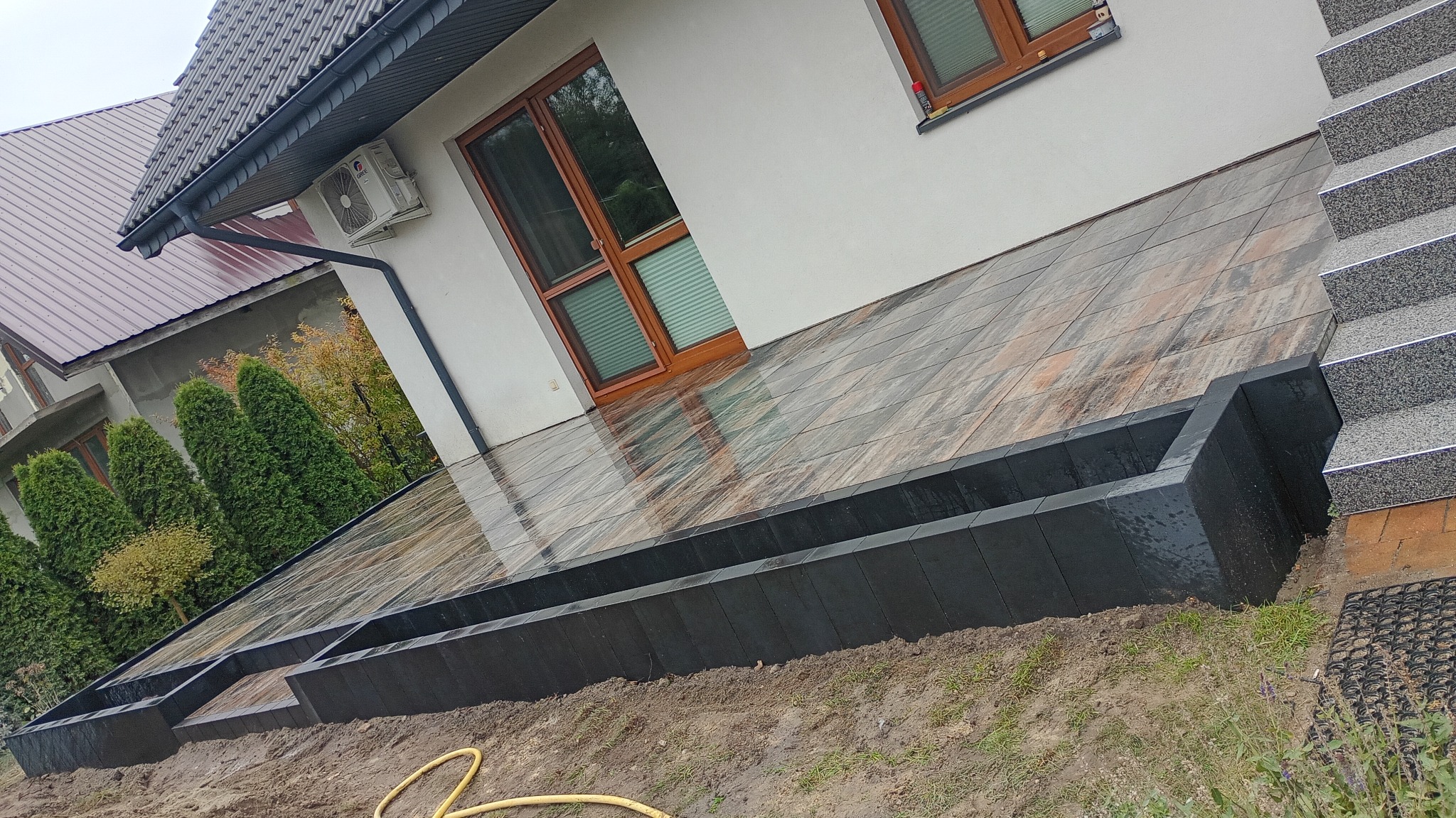 Nowoczesny taras z drewnopodobnej kostki, obrzeża z czarnych elementów, schody z szarego kamienia. Widok na dom z klimatyzatorem i brązowymi oknami.