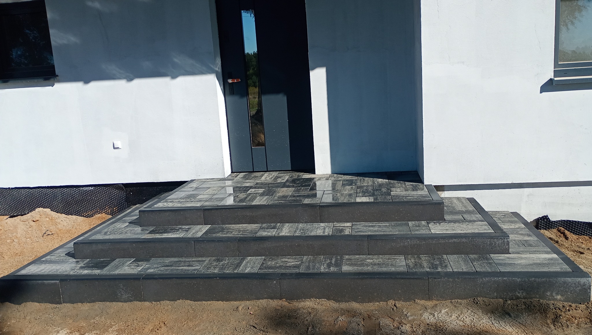 Schody wejściowe z kostki brukowej w odcieniach szarości prowadzące do nowoczesnego domu z ciemnymi drzwiami. Widoczne detale wykończenia i otoczenie.