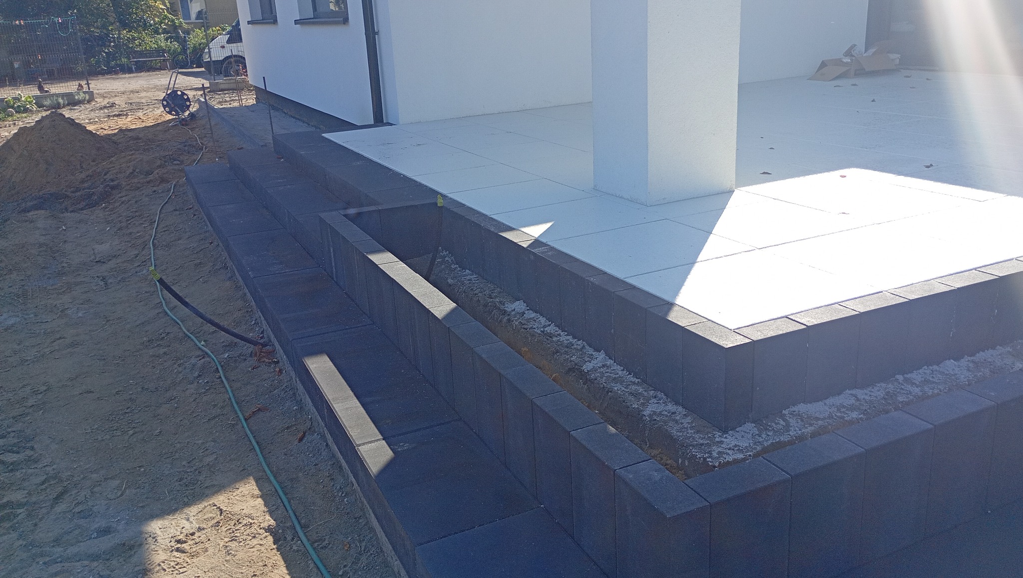 Schody z ciemnej kostki brukowej prowadzące do jasnego tarasu. Widoczny fragment domu i teren przygotowany pod dalsze prace. Oświetlenie słoneczne.