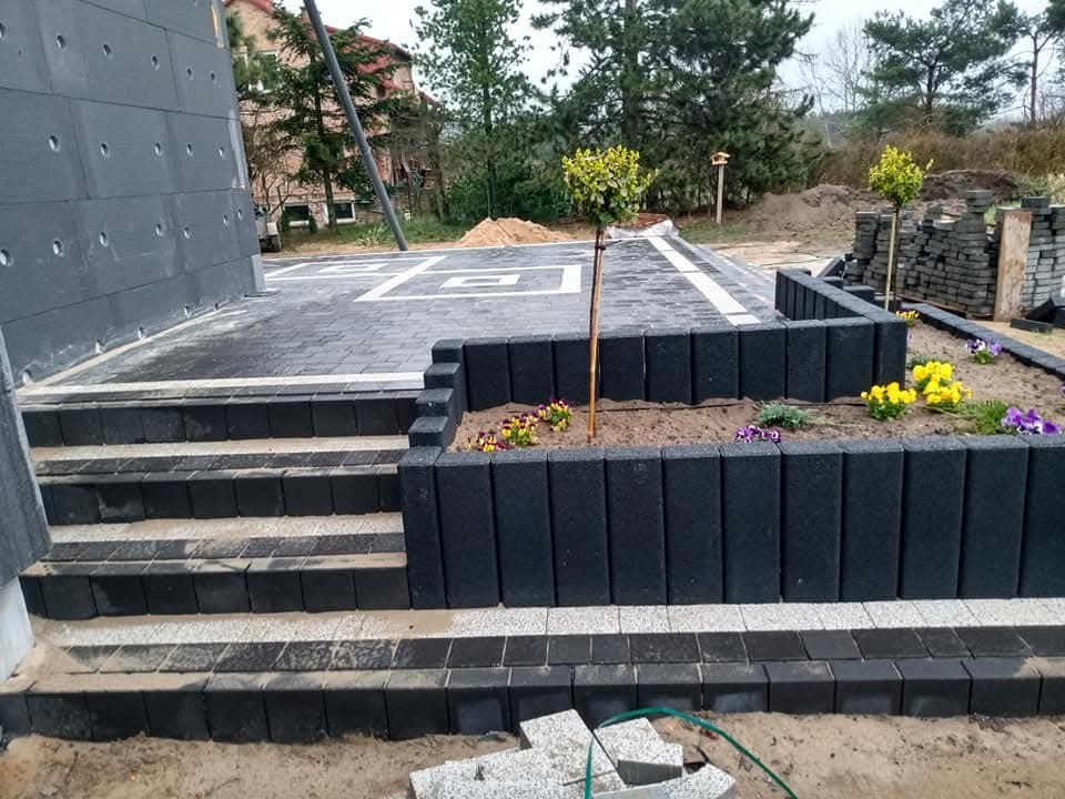 Schody i taras z kostki brukowej w odcieniach szarości, obrzeża z czarnych bloczków, rabaty z kwiatami. Widoczne elementy architektury budynku i zieleni.