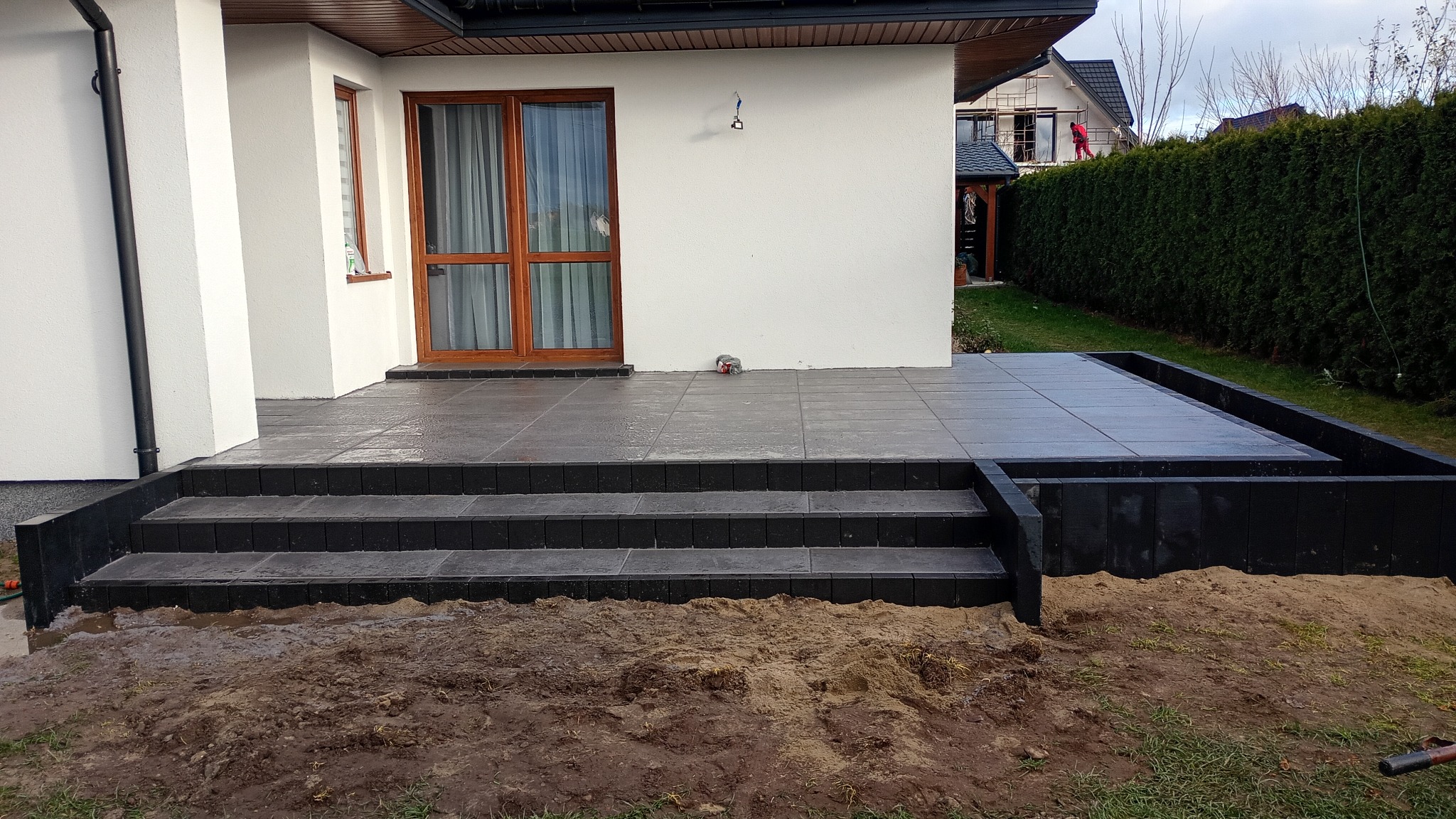 Nowoczesny taras z ciemnej kostki brukowej, schody wejściowe i obrzeże rabaty. Widok na biały dom z drewnianą stolarką okienną i zielony żywopłot w tle.