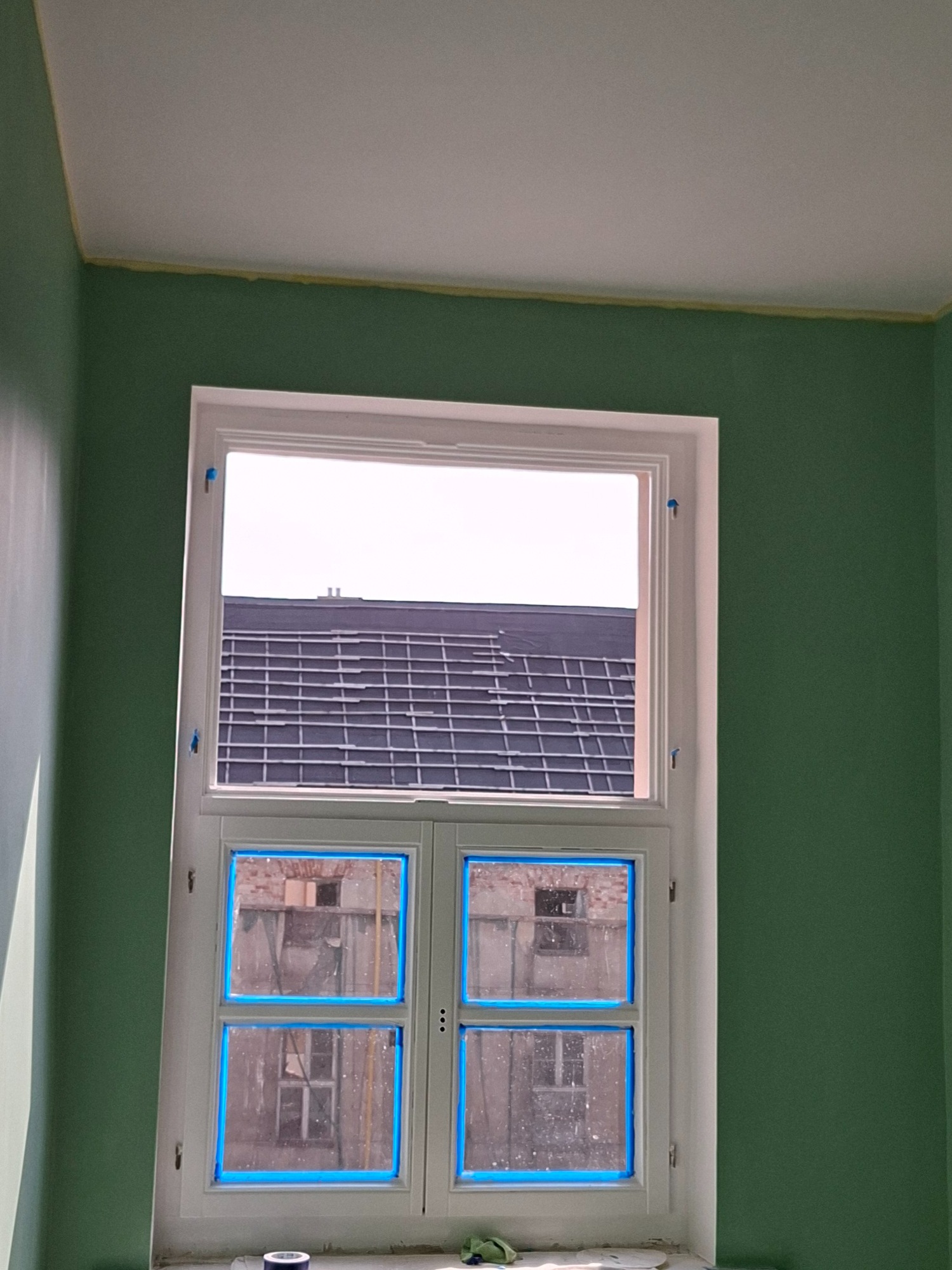 Pokój w trakcie malowania: ściany w odcieniach zieleni, okno zabezpieczone niebieską taśmą malarską, sufit biały z żółtą taśmą przy krawędzi. Widok na dach budynku.