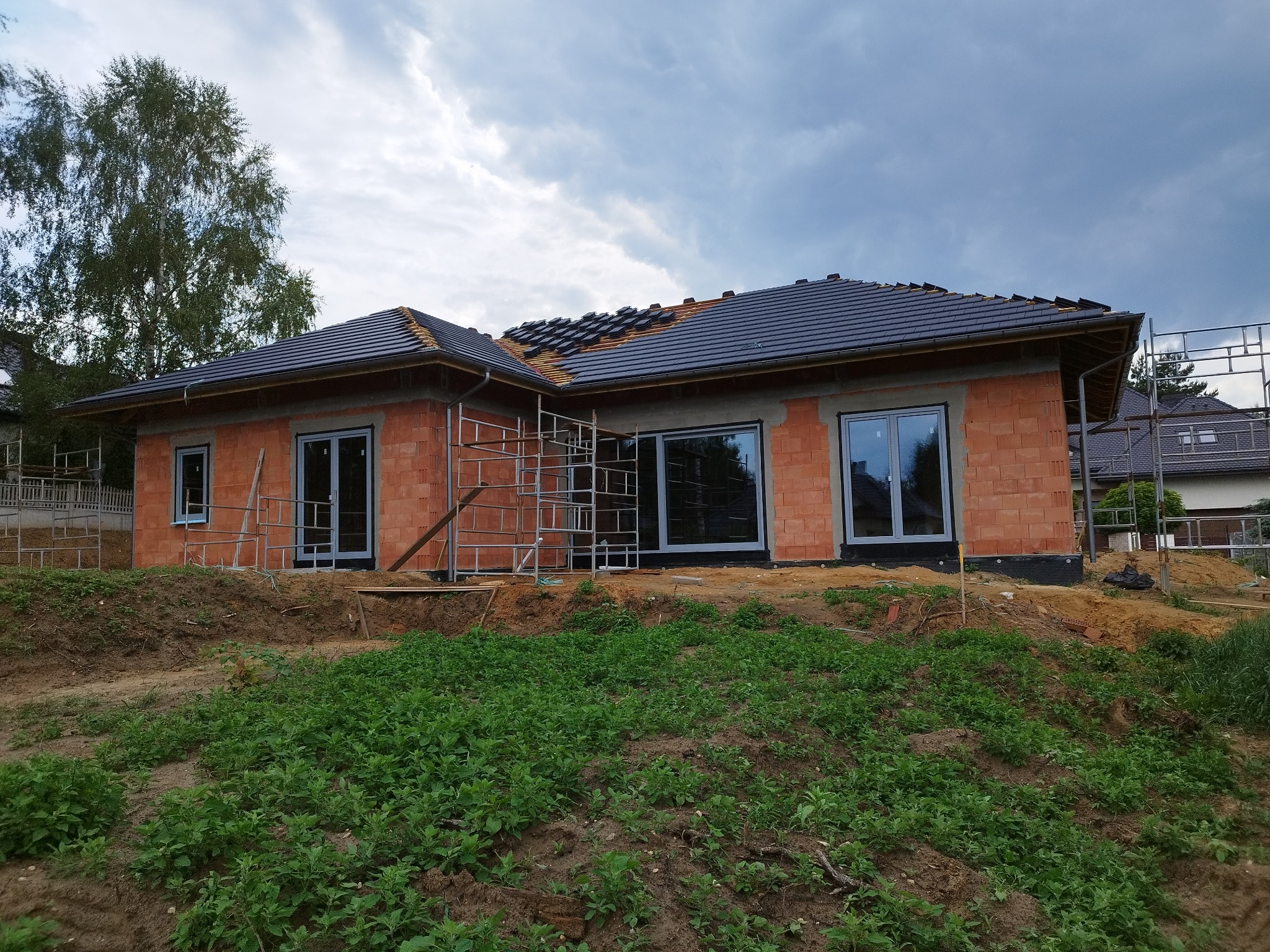 Dom w trakcie budowy z częściowo ułożonym dachem z dachówki, widoczne okna z szarymi ramami i rusztowania. Budynek z czerwonej cegły.