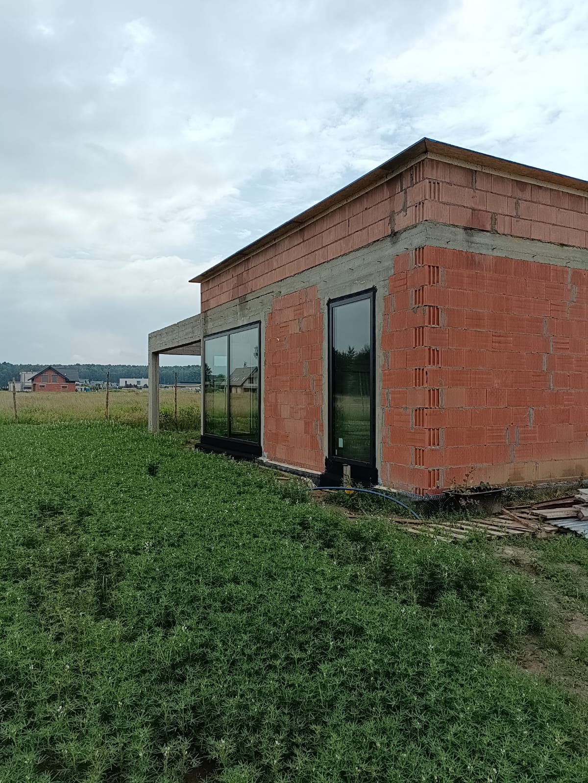 Surowy budynek z czerwonej cegły z zamontowanymi czarnymi oknami, widok z zewnątrz na tle zieleni i pochmurnego nieba. Etap budowy.