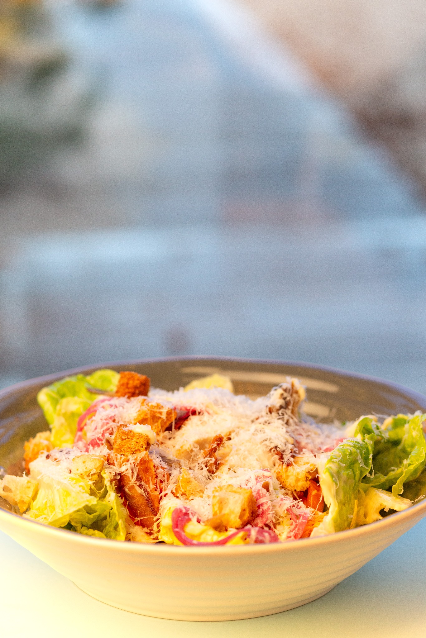 Sałatka cezar w szarej misce, z widocznymi grzankami, serem Grana Padano i czerwoną cebulą, na rozmytym tle.