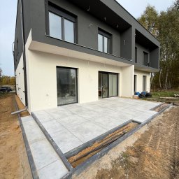 VR-BUD - Nowoczesny dom z szarą elewacją i tarasem wyłożonym jasnymi płytami. Widoczne przygotowania do wykończenia obrzeży tarasu. Prace ziemne w toku.