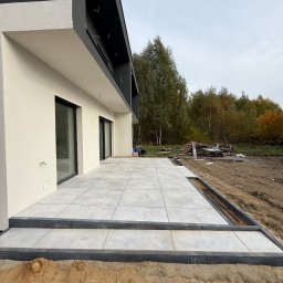 VR-BUD - Nowoczesny taras z szarych płyt, wykończony czarną obwódką, przylegający do budynku z białymi ścianami i czarnym dachem. Widoczne przygotowania do zagospodarowania terenu.