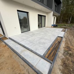 VR-BUD - Nowoczesny taras z szarych płyt, obrzeża z ciemnej kostki. Widoczne przygotowania do dalszych prac wykończeniowych wokół tarasu. Dom z szarą elewacją w tle.