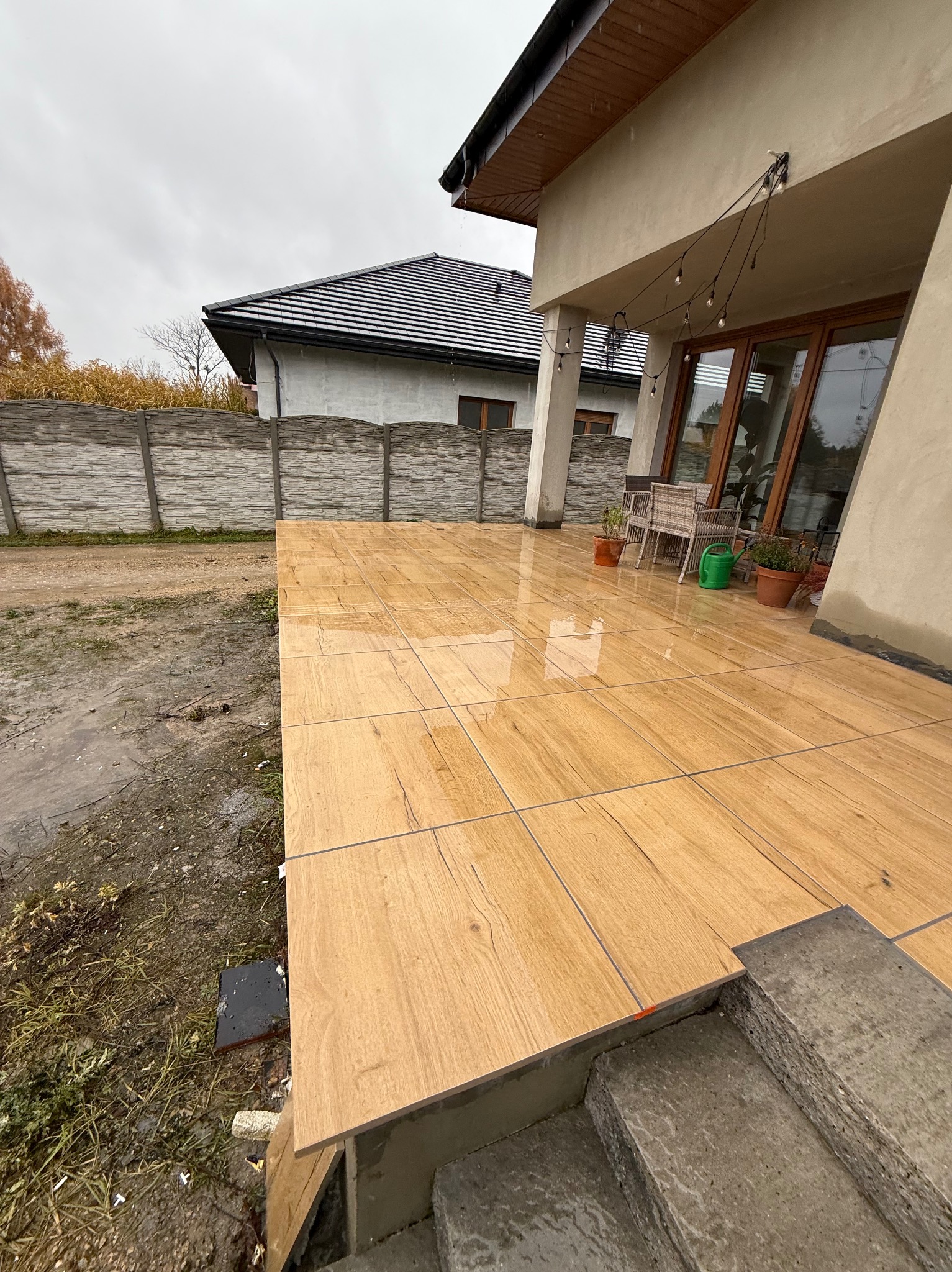 Wyłożony płytkami taras z drewnopodobnym wzorem, widok z boku na schody wejściowe. W tle dom z oknami i dekoracyjnymi lampkami, oraz ogrodzenie.