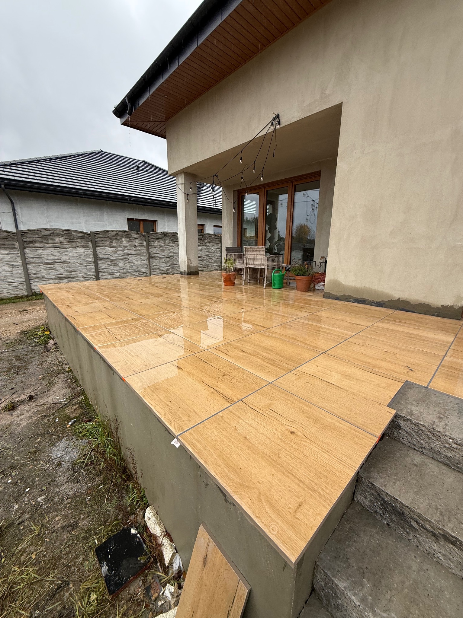 Wykończony taras z płytek imitujących drewno, z widocznymi fugami i schodami. Częściowo widoczne narzędzia i materiały budowlane. Tło stanowi elewacja budynku.