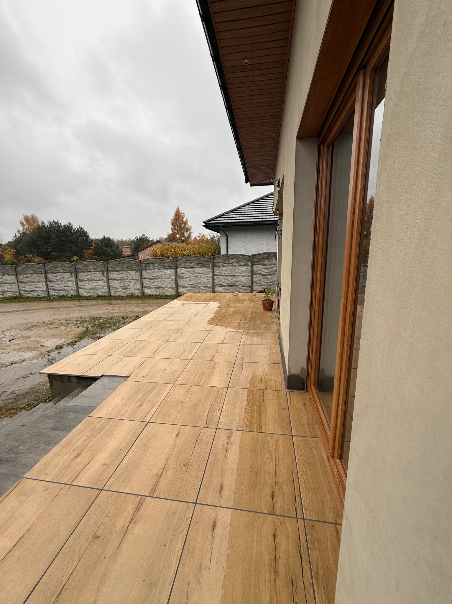 Kafelkowany taras zewnętrzny z drewnopodobnymi płytkami, częściowo ukończony, z widocznym fragmentem domu i ogrodu w tle, w pochmurny dzień.