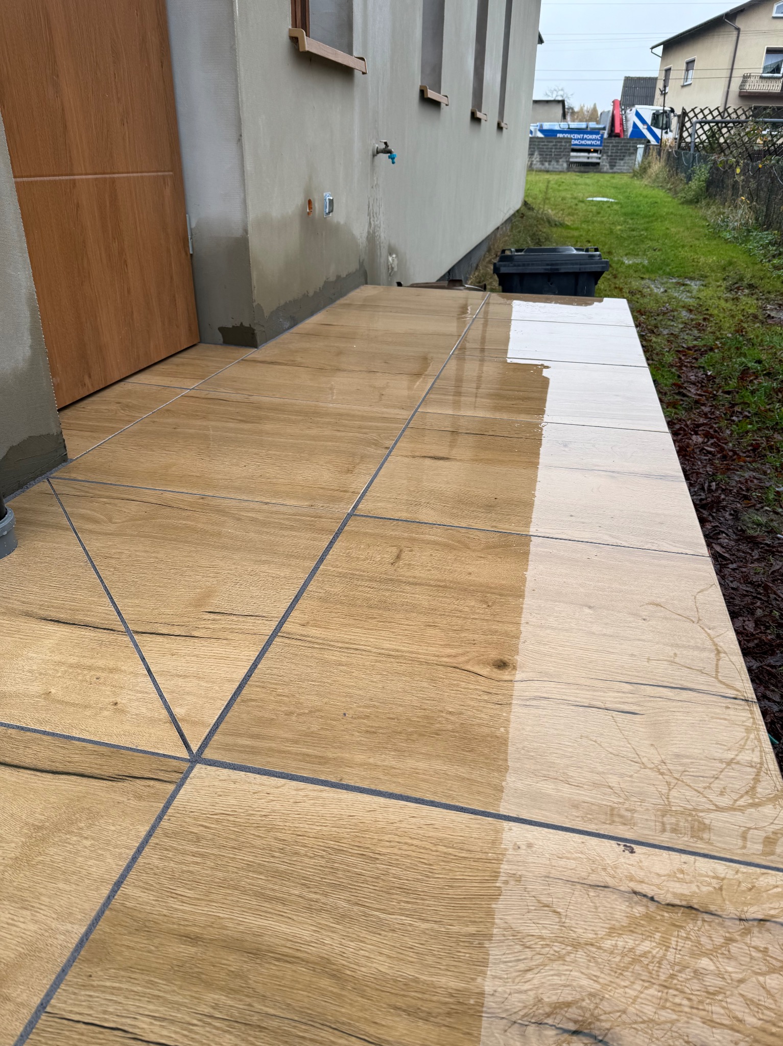 Ułożone płytki imitujące drewno na zewnątrz budynku, częściowo mokre, z widocznymi fugami i fragmentem elewacji. Widok na świeżo wykończony taras.