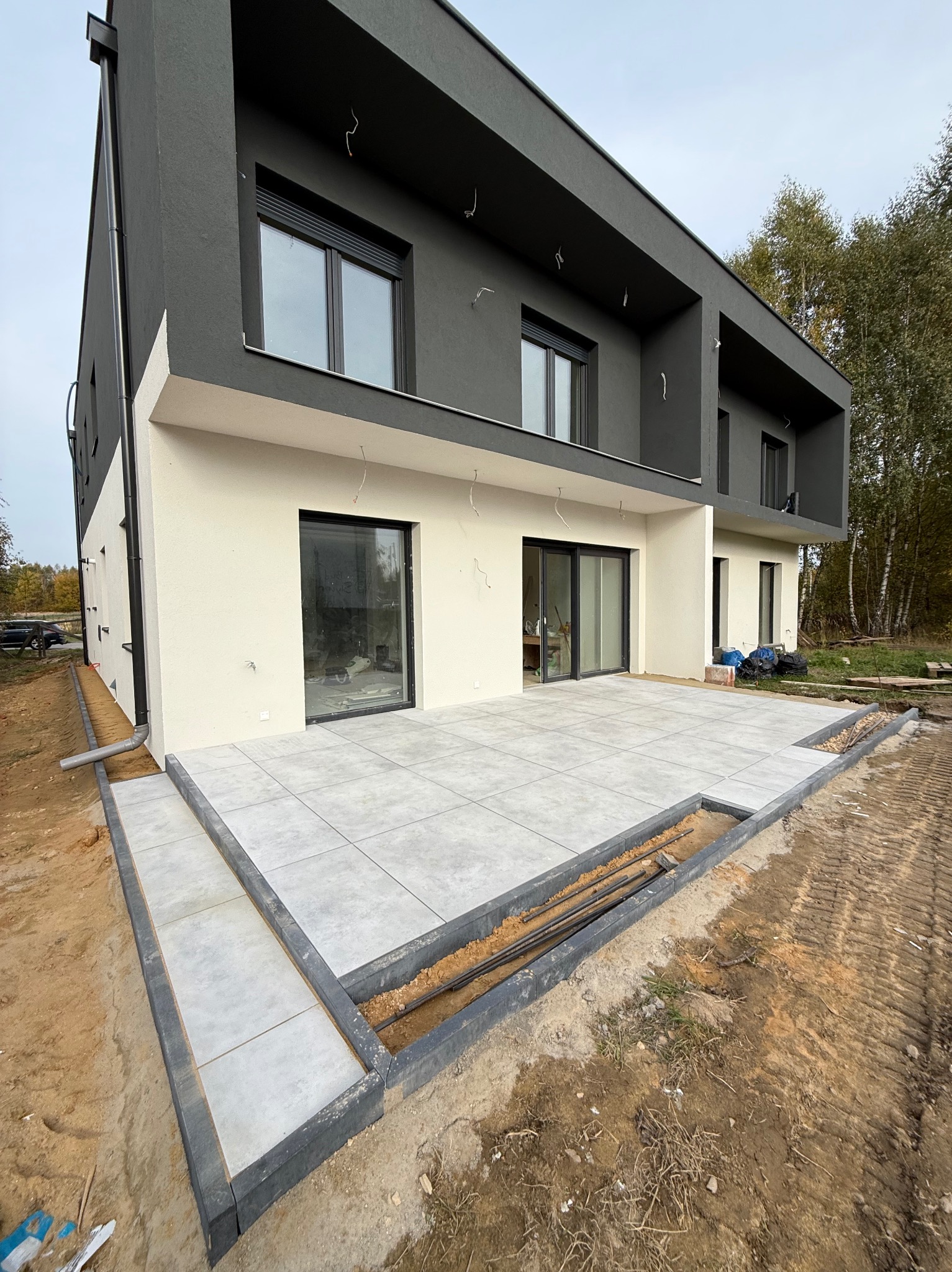 Nowoczesny dom z szarą elewacją i tarasem wyłożonym jasnymi płytami. Widoczne przygotowania do wykończenia obrzeży tarasu. Prace ziemne w toku.