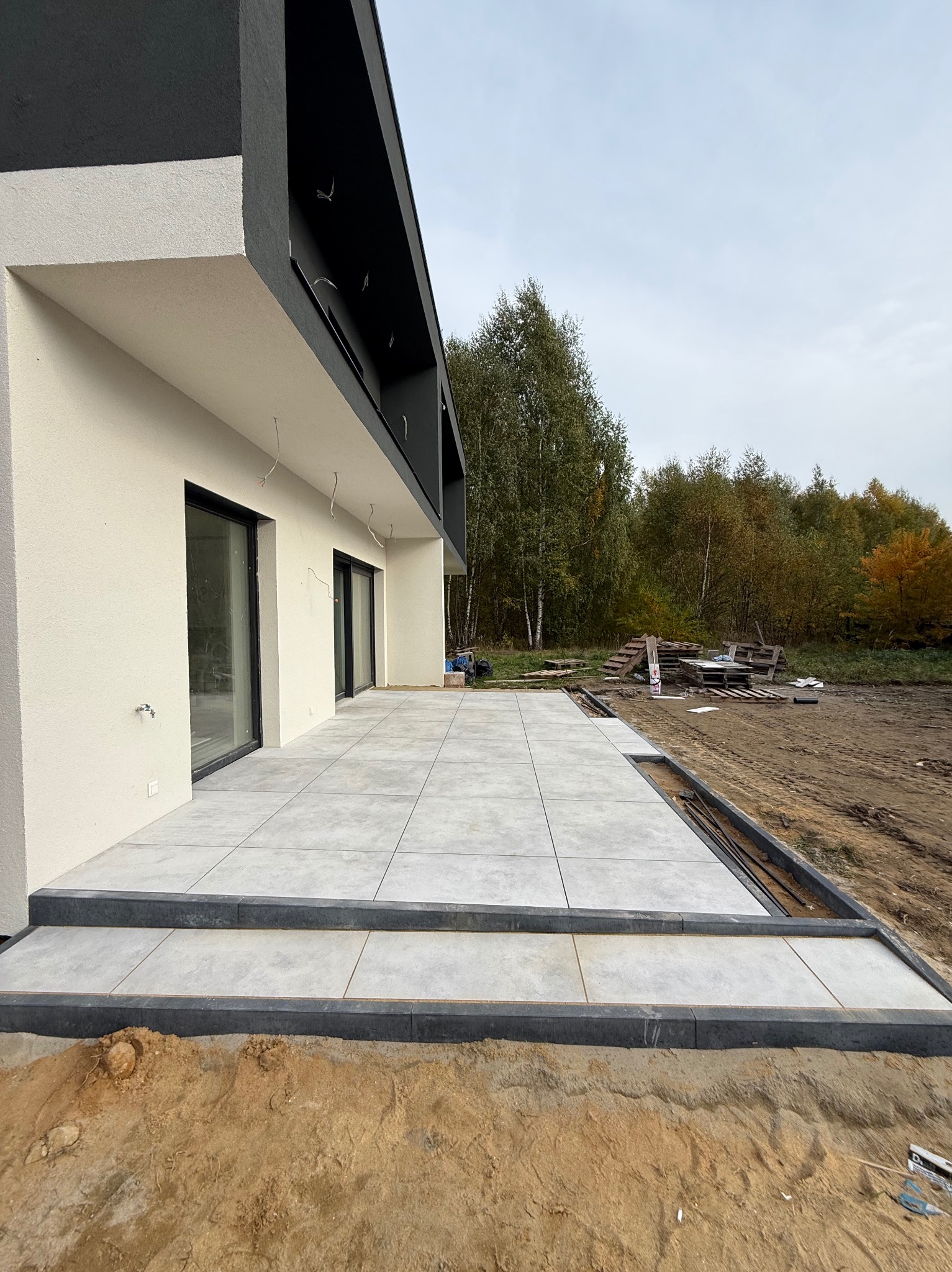 Nowoczesny taras z szarych płyt, wykończony czarną obwódką, przylegający do budynku z białymi ścianami i czarnym dachem. Widoczne przygotowania do zagospodarowania terenu.