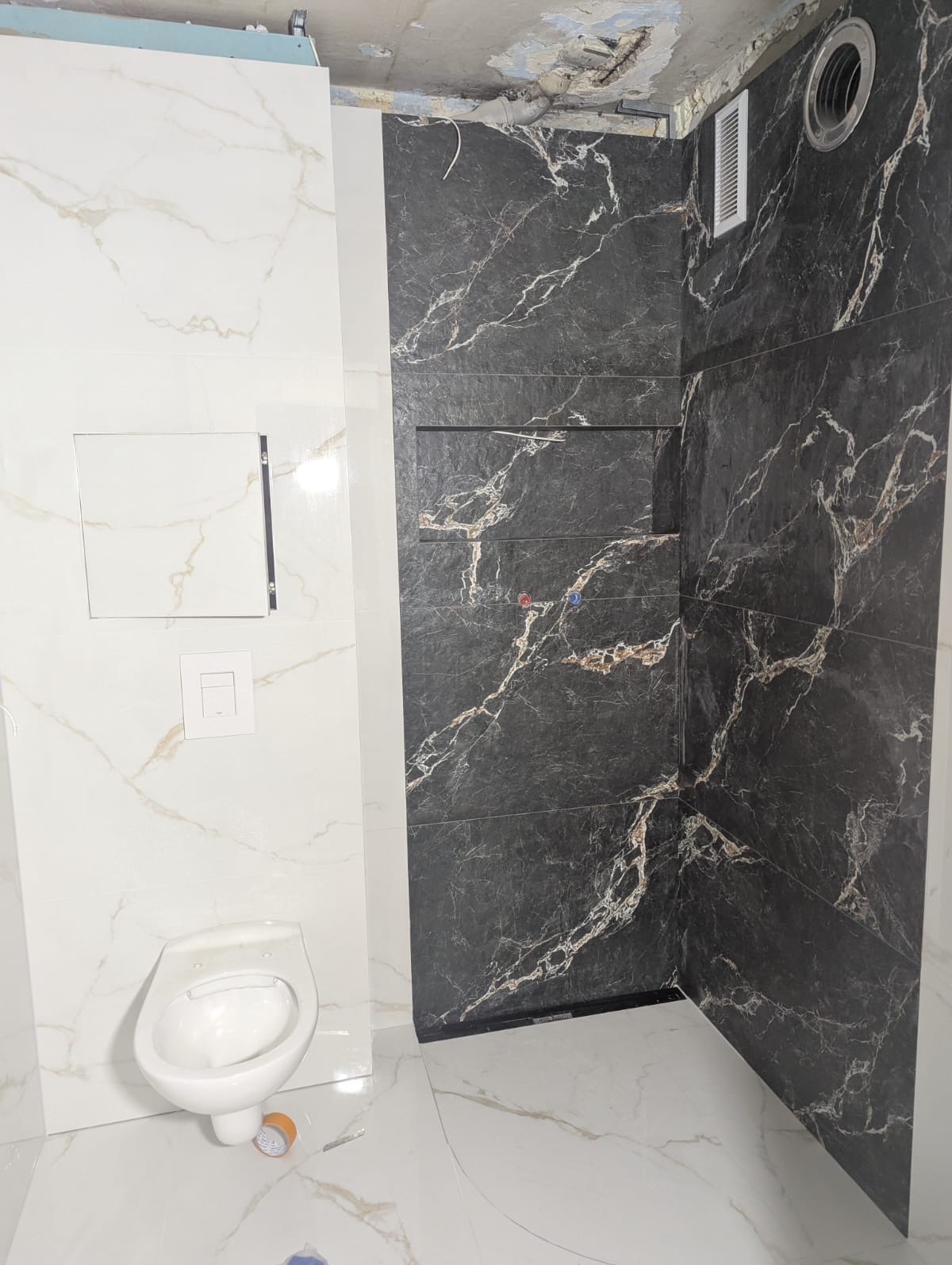 Nowoczesna łazienka po remoncie: białe i czarne marmurowe płytki, podwieszana toaleta, wnęki w strefie prysznica. Widoczne niedociągnięcia na suficie.