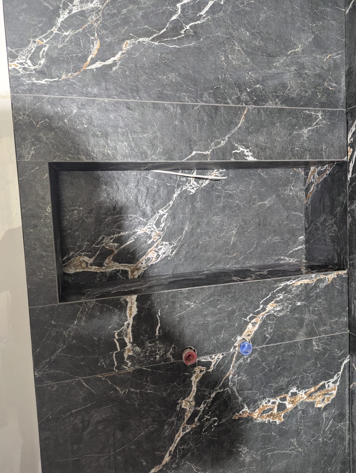 Ciemne, marmurowe płytki w łazience z wnęką, widoczne przyłącza wodne. Minimalistyczny design z wyraźnym wzorem żyłkowania. Wykończenie ściany w nowoczesnym stylu.