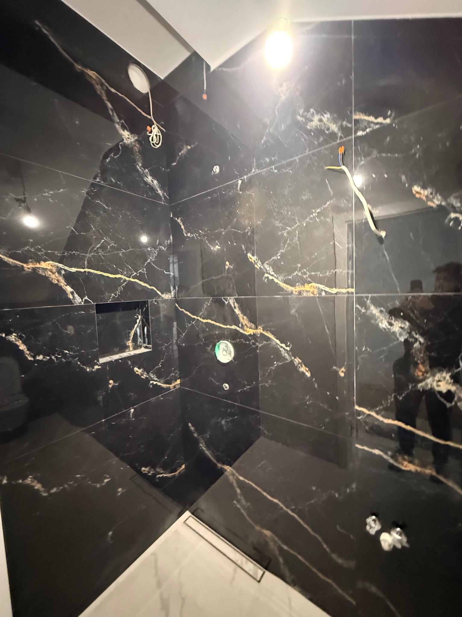Czarne płytki imitujące marmur z złotymi żyłami w nowoczesnej kabinie prysznicowej z odpływem liniowym i wnęką. Widoczne elementy instalacji elektrycznej.