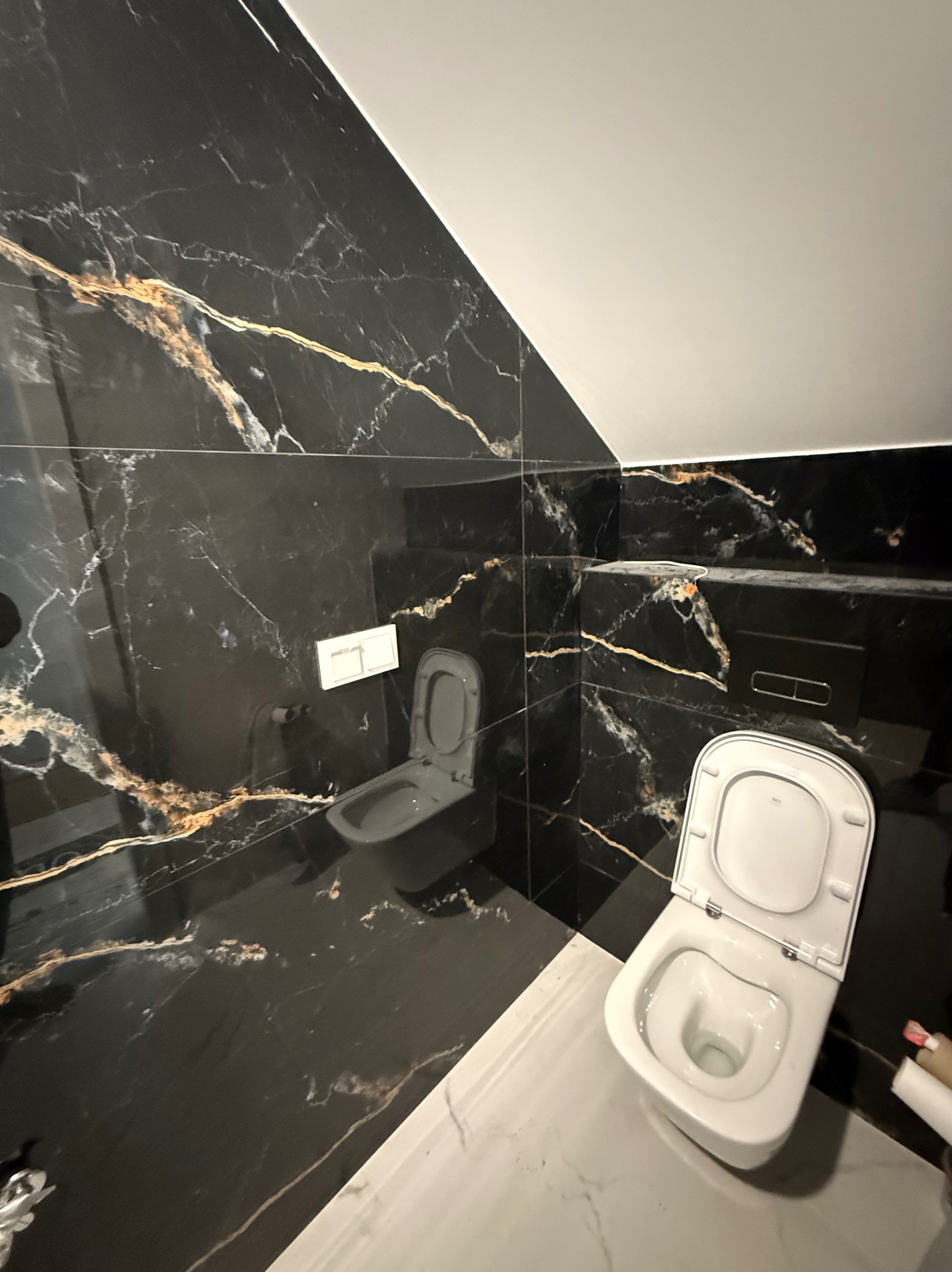 Elegancka łazienka z czarnymi płytkami imitującymi marmur ze złotymi żyłami, wisząca toaleta i biała podłoga. Ukośny sufit dodaje unikalnego charakteru.