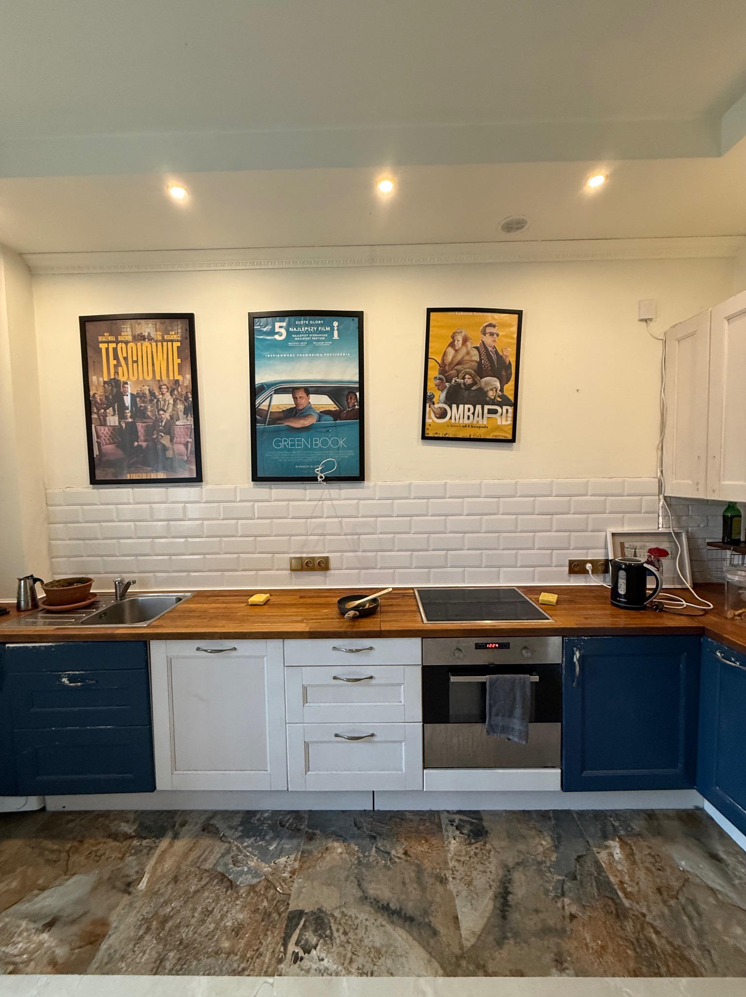 Kuchnia po remoncie: białe płytki na ścianie, drewniany blat, niebieskie i białe szafki. Na ścianie wiszą plakaty filmowe w czarnych ramkach. Nowoczesny design.