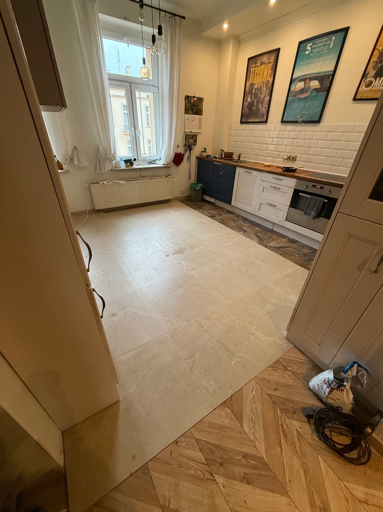 Jasna kuchnia po remoncie: beżowe płytki podłogowe imitujące marmur przechodzą w parkiet ułożony w jodełkę. Widoczne białe meble kuchenne i plakaty na ścianie.