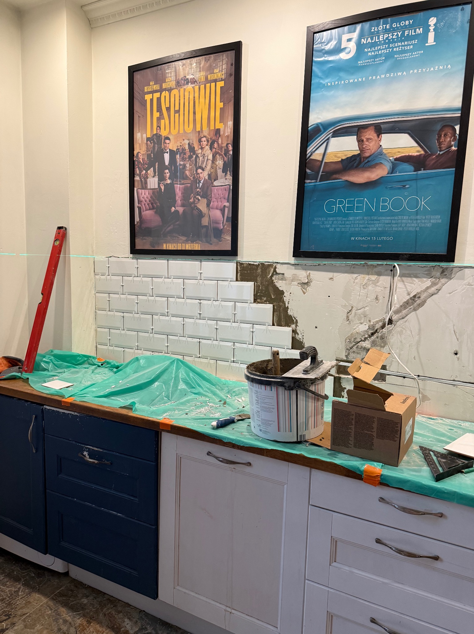 Układanie białych płytek na ścianie w kuchni, widoczne narzędzia glazurnicze, poziomica laserowa, plakaty filmowe na ścianie. Etap prac wykończeniowych.