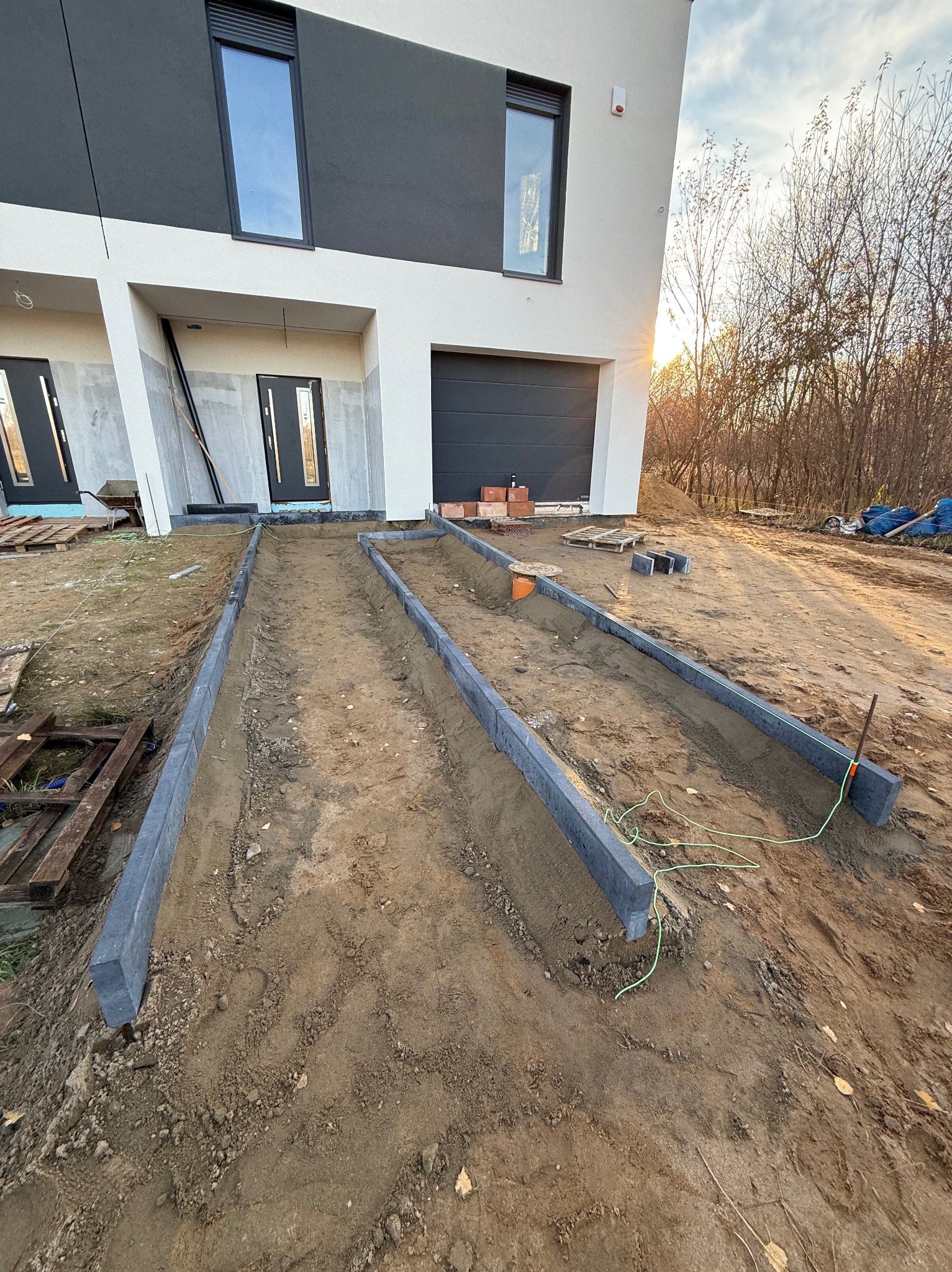 Montaż obrzeży chodnikowych z szarej kostki przy nowoczesnym domu z garażem. Widoczne przygotowane podłoże i narzędzia. Prace brukarskie w toku, ciepłe światło zachodzącego słońca.