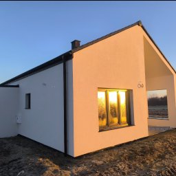 Usługi Ogolnobudowlane - Nowoczesny, parterowy dom z białymi ścianami i czarnym dachem w promieniach zachodzącego słońca, z widocznym numerem domu '60'.