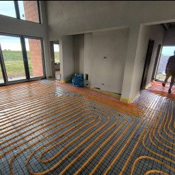 Usługi Ogolnobudowlane - Rozłożona instalacja ogrzewania podłogowego w nowym domu z dużymi oknami. Pomarańczowe rurki ułożone na czarnej siatce, widoczny fragment krajobrazu za oknem.