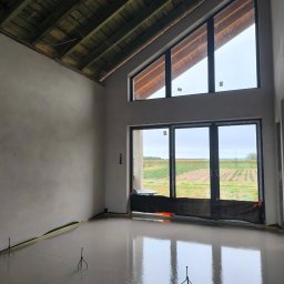 Usługi Ogolnobudowlane - Wnętrze domu w trakcie budowy: świeżo wylana, mokra posadzka z widocznymi wspornikami, surowy strop drewniany i duże okna z widokiem na pole.
