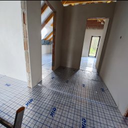 Usługi Ogolnobudowlane - Rozłożona folia izolacyjna z nadrukiem na podłodze w surowym wnętrzu domu w trakcie budowy. Widoczne ściany w stanie deweloperskim i fragment drabiny.