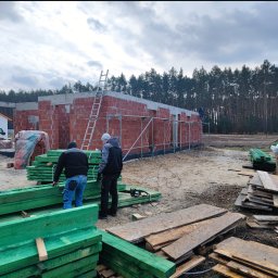 Usługi Ogolnobudowlane - Budowa domu w trakcie: ściany z czerwonej cegły, rusztowania, drabina. Dwóch robotników przy zielonych belkach. W tle las i fragment drugiego budynku.