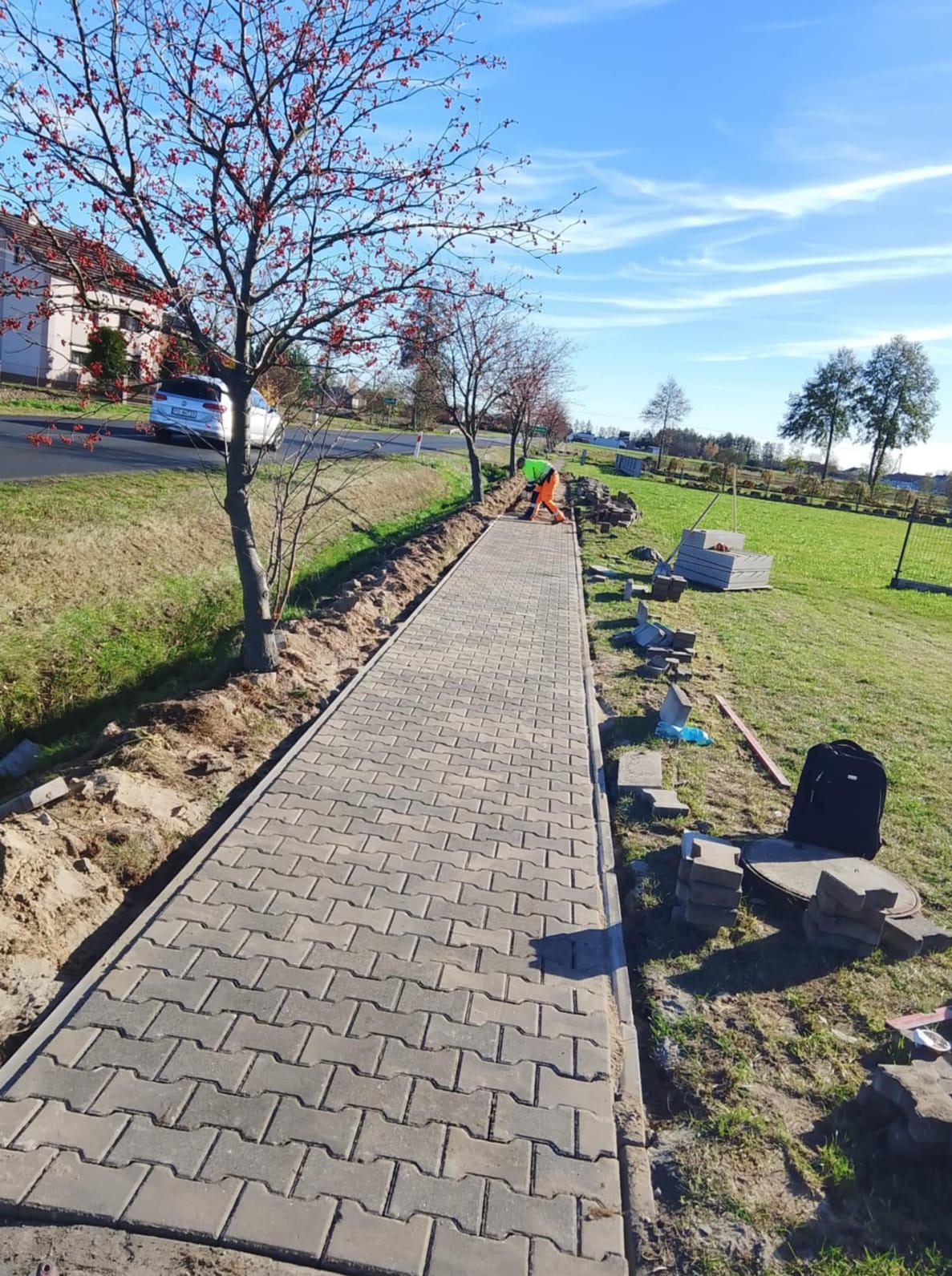 Długa ścieżka z szarej kostki brukowej w trakcie układania, widoczny pracownik w odblaskowej kamizelce, narzędzia i materiały budowlane. Słoneczny dzień.
