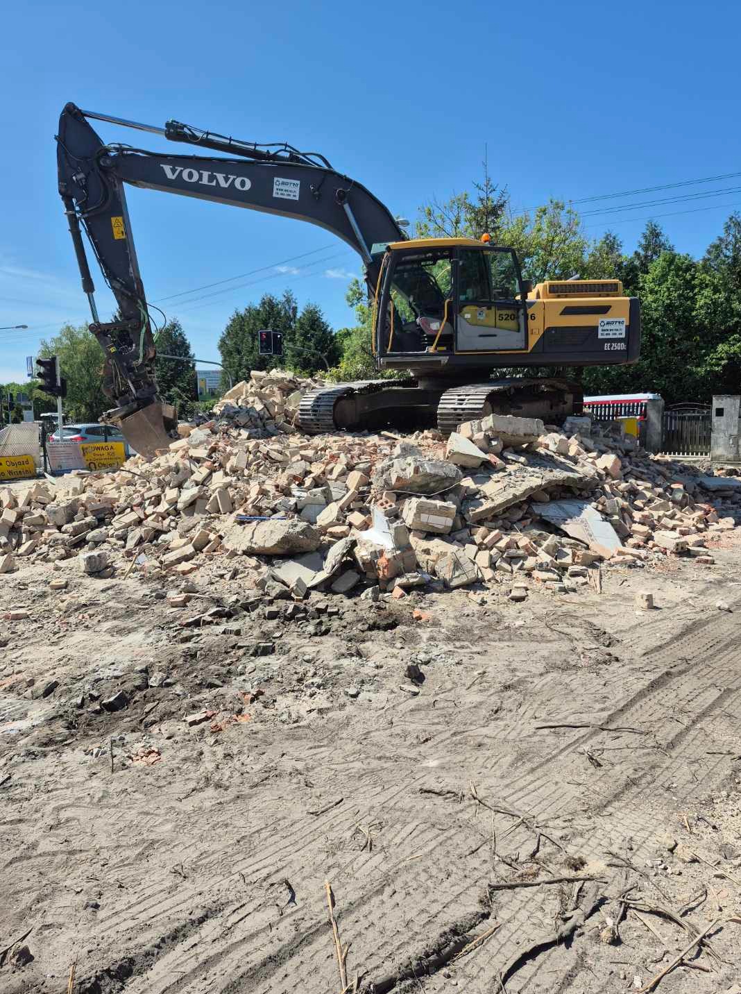 Żółto-szara koparka Volvo na stercie gruzu ceglanego po rozbiórce, na tle zieleni i błękitnego nieba. Widoczne ślady opon na ziemi.