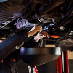 MasterOil - Serwis Samochodowy - Mechanik odkręca śrubę pod samochodem, widoczna misa na olej i podnośnik. Ujęcie z dołu pojazdu.