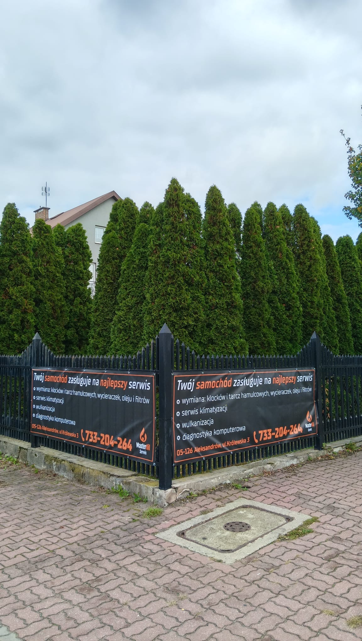 Ogrodzenie warsztatu z banerem reklamującym wymianę klocków, serwis klimatyzacji, wulkanizację i diagnostykę komputerową. Tło z zielonych drzew.