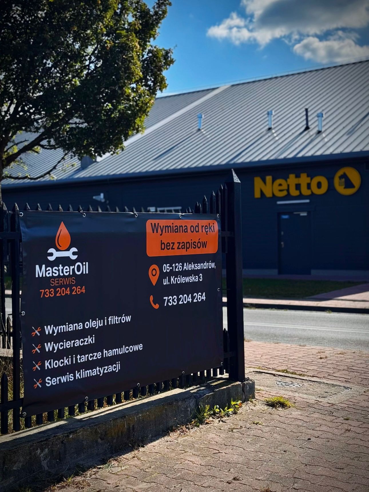Baner reklamowy warsztatu samochodowego MasterOil z ofertą wymiany oleju, filtrów, wycieraczek, klocków hamulcowych i serwisu klimatyzacji, umieszczony przed budynkiem Netto.