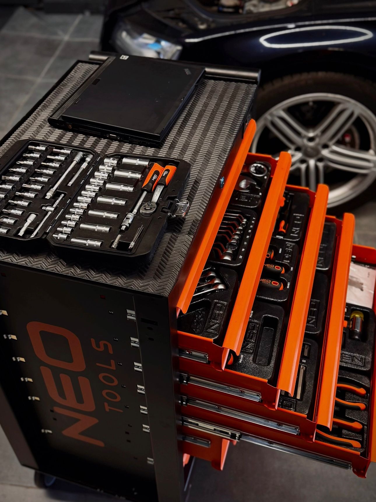 Wózek warsztatowy Neo Tools z otwartymi szufladami, pełnymi narzędzi. W tle widoczny fragment samochodu z otwartą maską i koło.