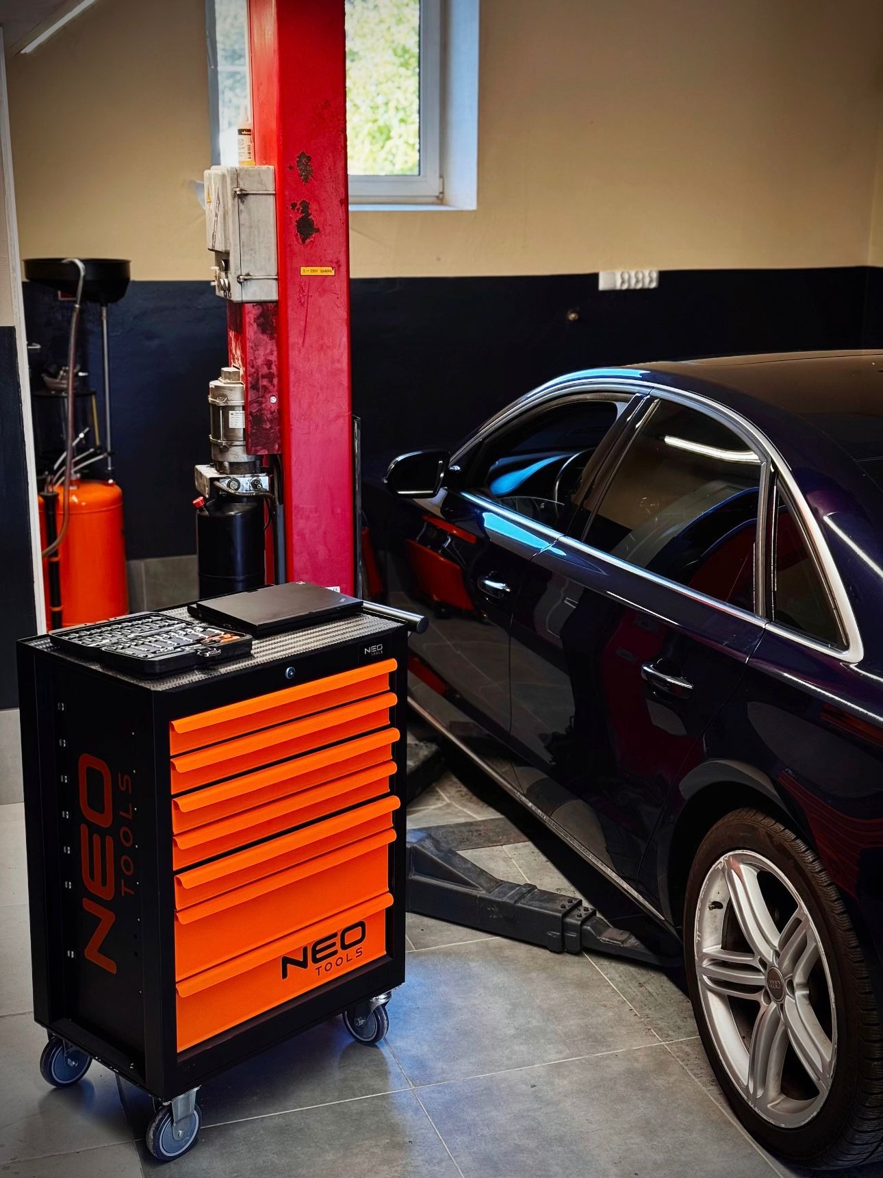 Samochód na podnośniku w warsztacie, obok wózek narzędziowy Neo Tools. Widoczny czerwony słup podnośnika i okno w tle. Wnętrze warsztatu z ciemnymi elementami.