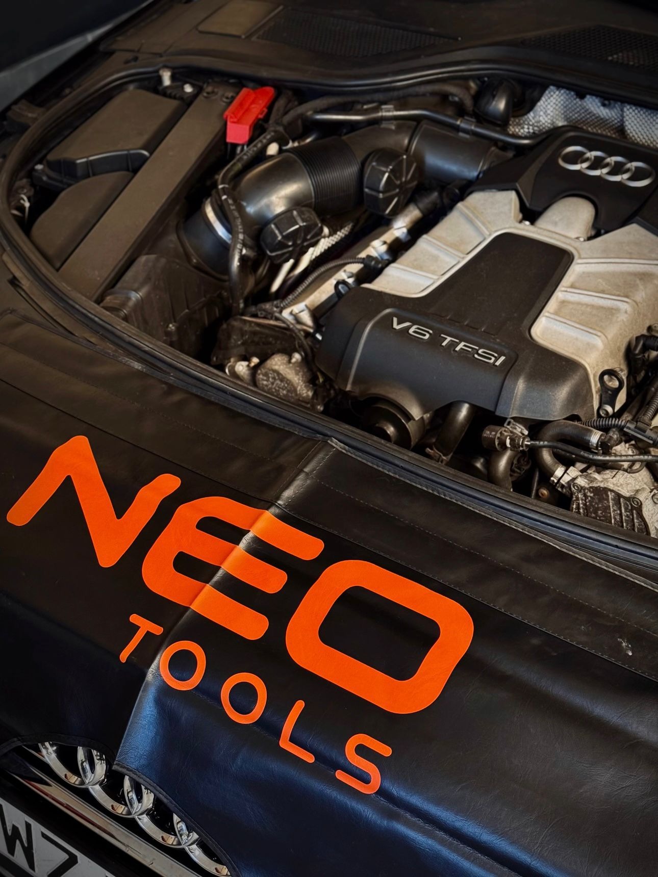 Otwarta maska Audi z widocznym silnikiem V6 TFSI, zabezpieczona czarną osłoną z pomarańczowym logo NEO Tools. Widoczna bateria i elementy instalacji.