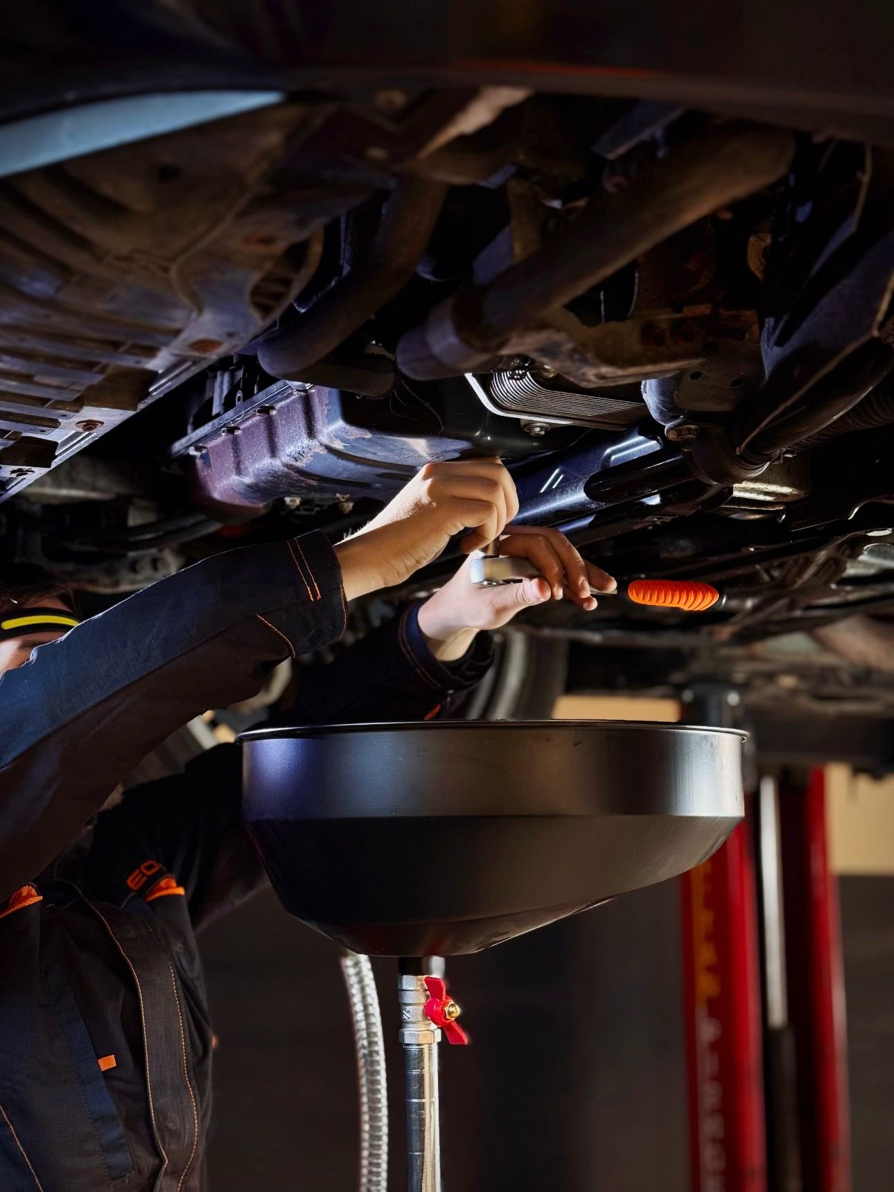 Mechanik odkręca śrubę pod samochodem, widoczna misa na olej i podnośnik. Ujęcie z dołu pojazdu.