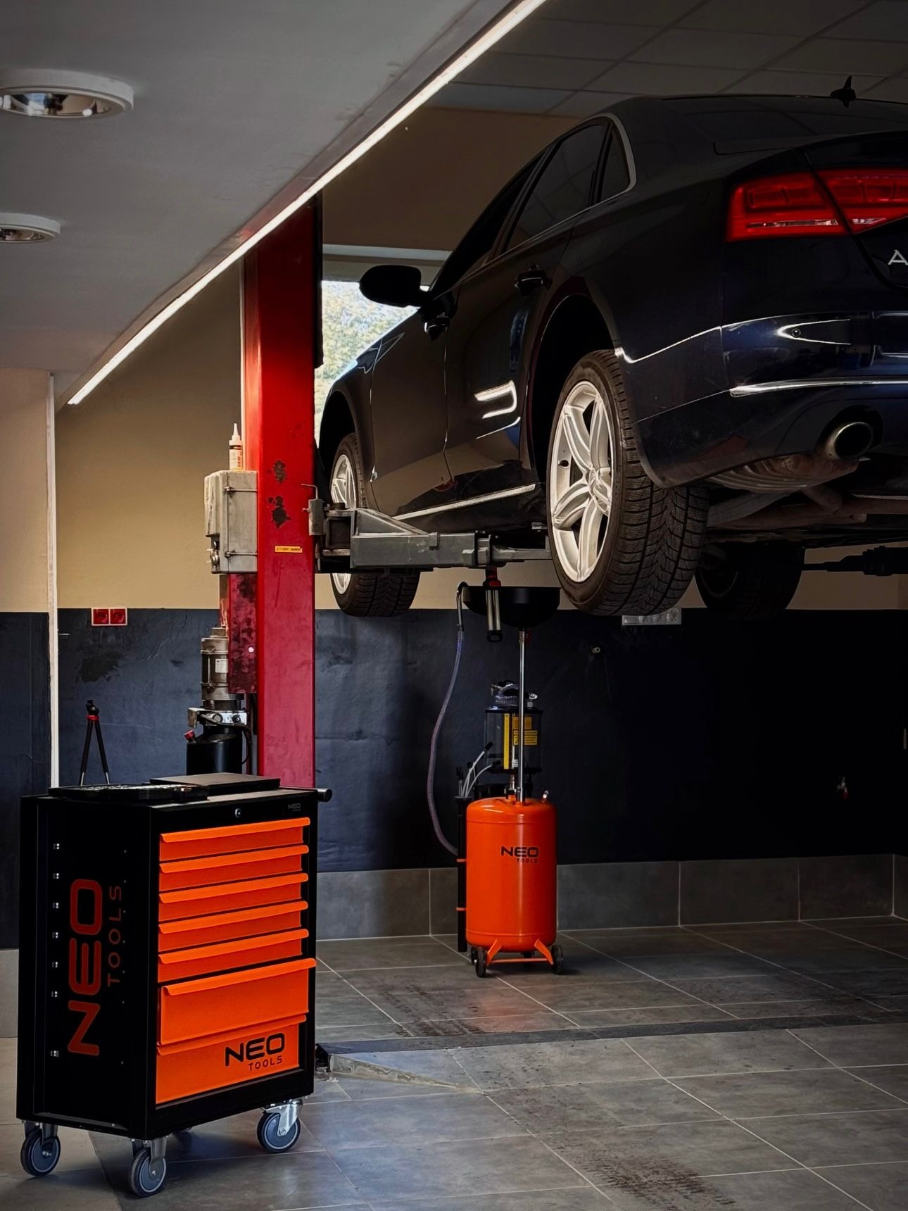 Ciemny sedan Audi na podnośniku w warsztacie. Widoczny wózek narzędziowy Neo Tools i pojemnik na zużyty olej. Oświetlenie LED.