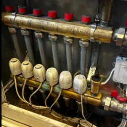 Rozdzielacz ogrzewania podłogowego z rurami i zaworami, opisany nazwami pomieszczeń (Salon, Kazimierz, Wiatr, Kuchnia). Widoczne narzędzia hydrauliczne.