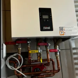 Biały piec gazowy Ferroli z miedzianą instalacją, zaworami (czerwony, żółty) i pompą obiegową. Widoczne płytki ceramiczne na ścianie i fragment grzejnika.